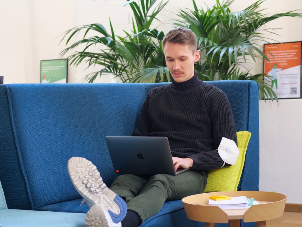 Person in black turtleneck working on laptop on blue sofa, with green plants in background and small table nearby.