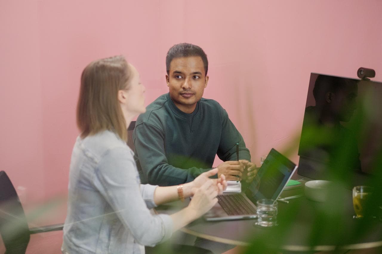Two colleagues in discussion at a desk with laptops, against a pink wall background in a modern office setting.
